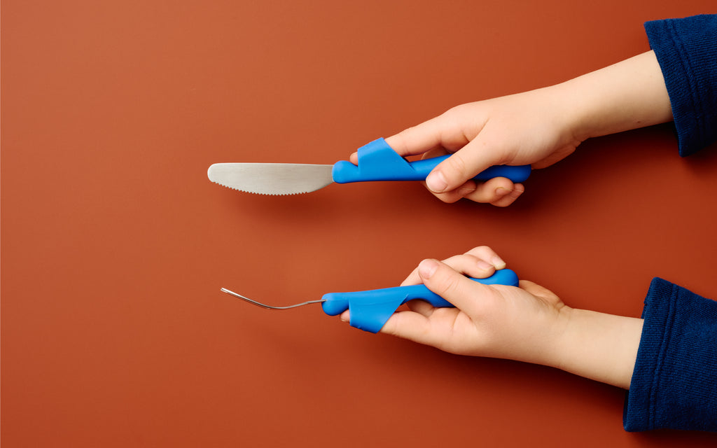 Children's hands holding Kinder Cutlery's blue training fork and knife set, which is designed to help them cut food independently, against a brown background.