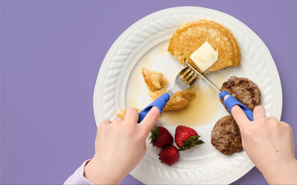 Children's hands using Kinder Cutlery's blue training fork and knife set to independently cut a pancake into bite-sized pieces on a white plate with breakfast sausage and strawberries against a purple background.